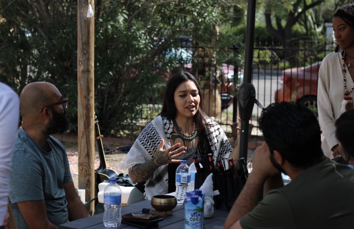 Ryanne speaking with kin gathered around a table outdoors, sharing a teaching with her hands