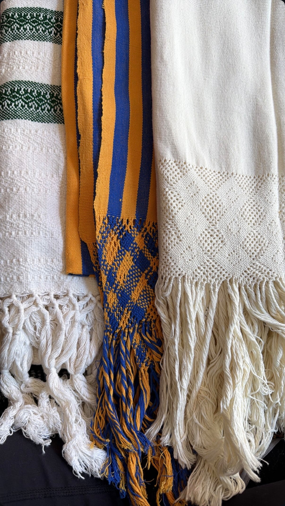 Handwoven rebozos with fringed ends — cream, green, and gold-and-blue cloths