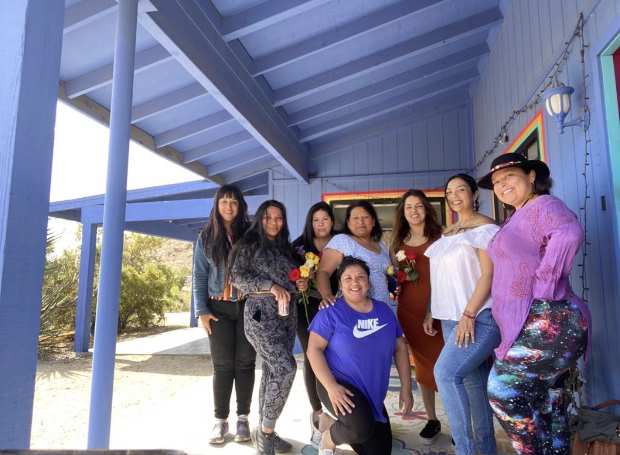 Group of women gathered under a blue porch in the desert, some holding roses